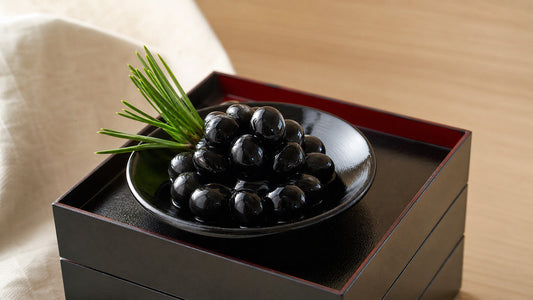 Glossy simmered kuromame black soybeans mounded in a black lacquer dish on a stacked Japanese New Year osechi jubako box, with a sprig of pine