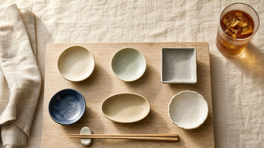 A Japanese snack board arranged and still empty: six small ceramic dishes on a pale wood board, iced barley tea alongside.