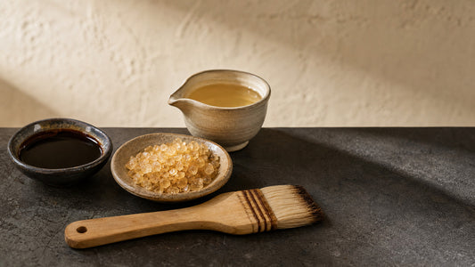 Tamari soy sauce in a dark ceramic bowl, coarse amber zarame sugar crystals in a shallow stoneware dish, and mirin in a pale spouted pitcher beside a flat hake brush, the ingredients of a Japanese teriyaki glaze
