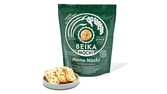 Green Beika mame mochi pouch standing on white beside a pale ceramic dish of bean-studded okaki rice crackers